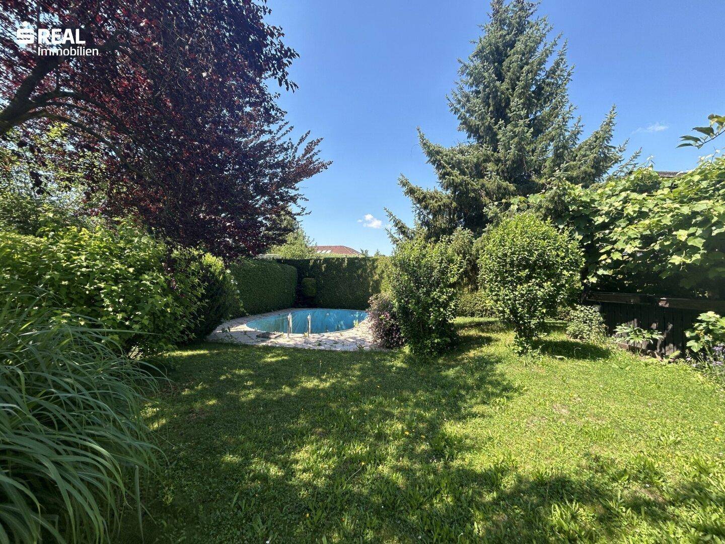 Gepflegter Gartenbereich mit kleinem Pool, umgeben von grünen Hecken und Bäumen unter blauem Himmel.