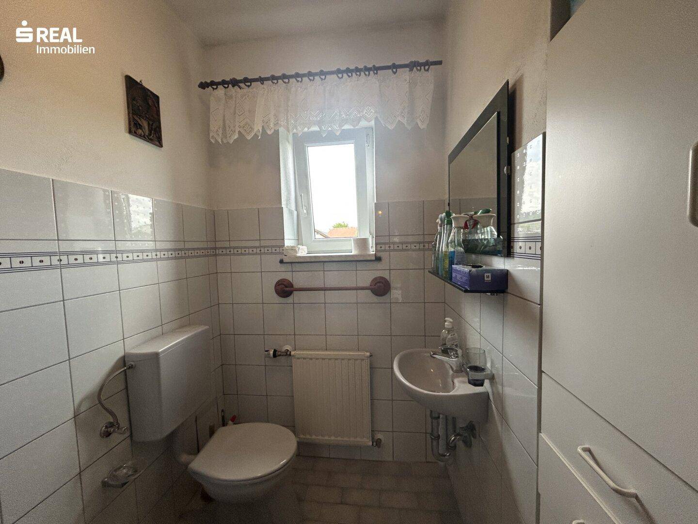 Kleines Gäste-WC mit weißen Fliesen, Toilette, Waschbecken und einem Fenster für Tageslicht.
