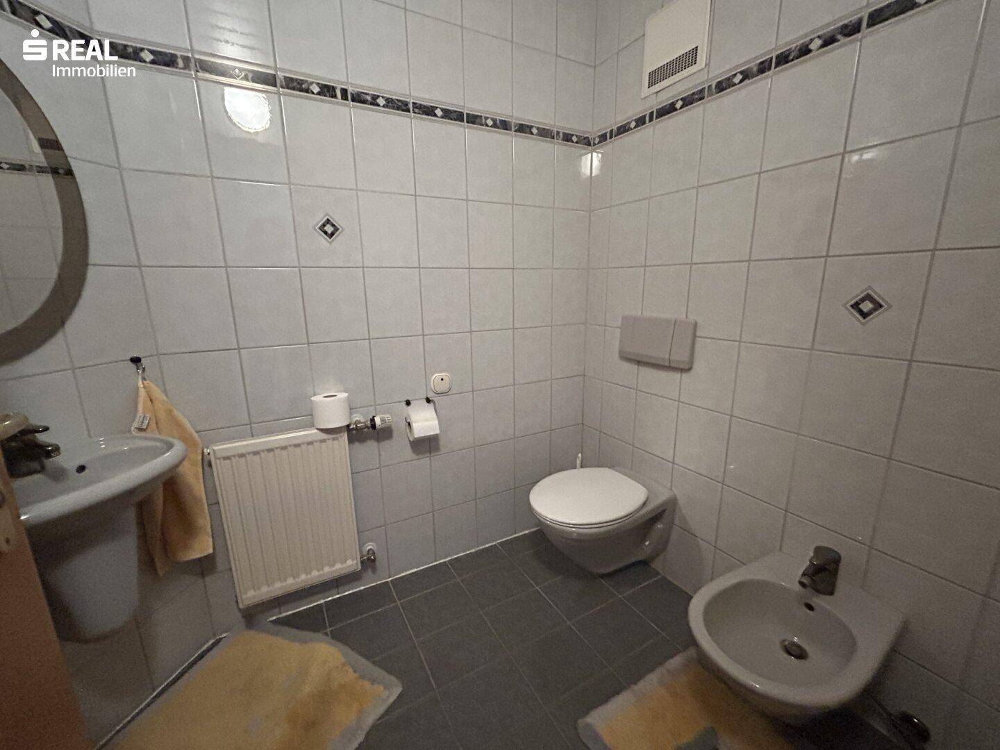 Badezimmer mit weißen und grauen Fliesen, ausgestattet mit Toilette, Bidet und Waschbecken.