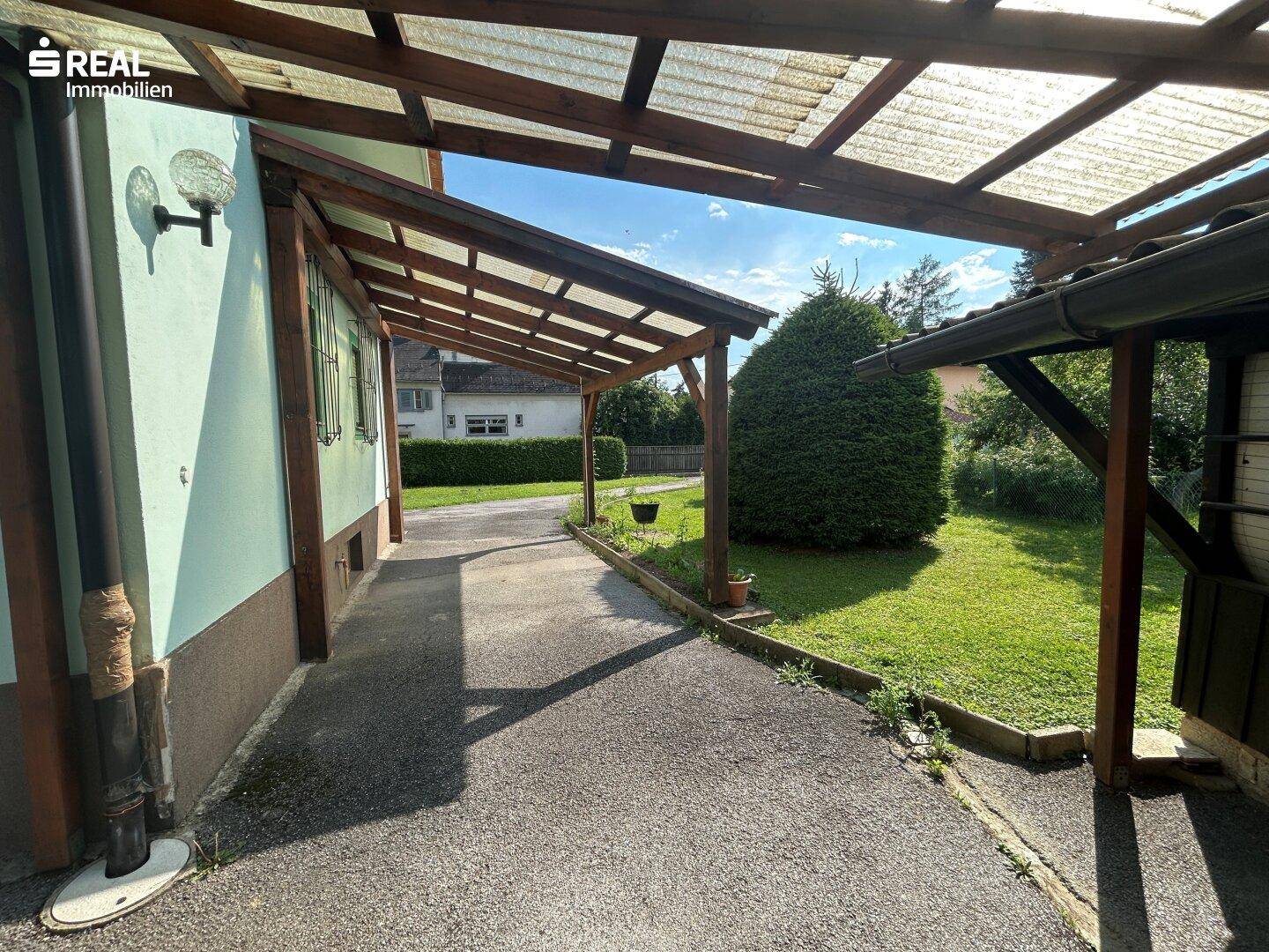 Überdachter Zugangsbereich neben dem Haus mit Holzbalken und Blick auf den Garten.