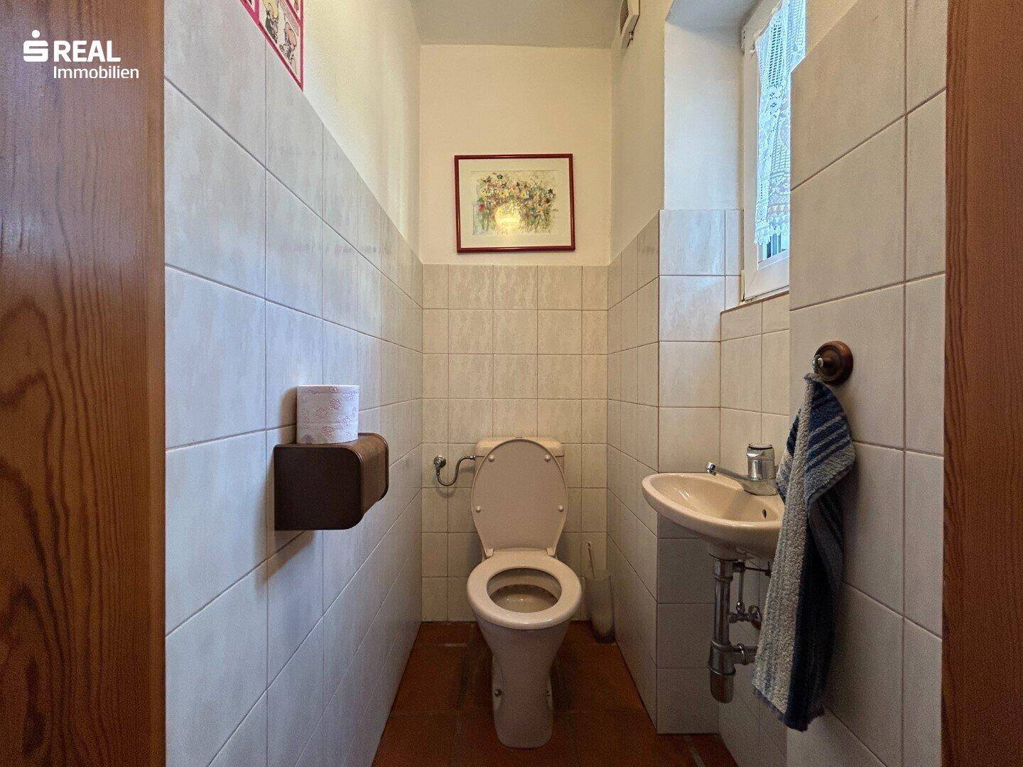 Kleines Gäste-WC mit weißen Fliesen, Toilette, Waschbecken und einem Fenster für Tageslicht.