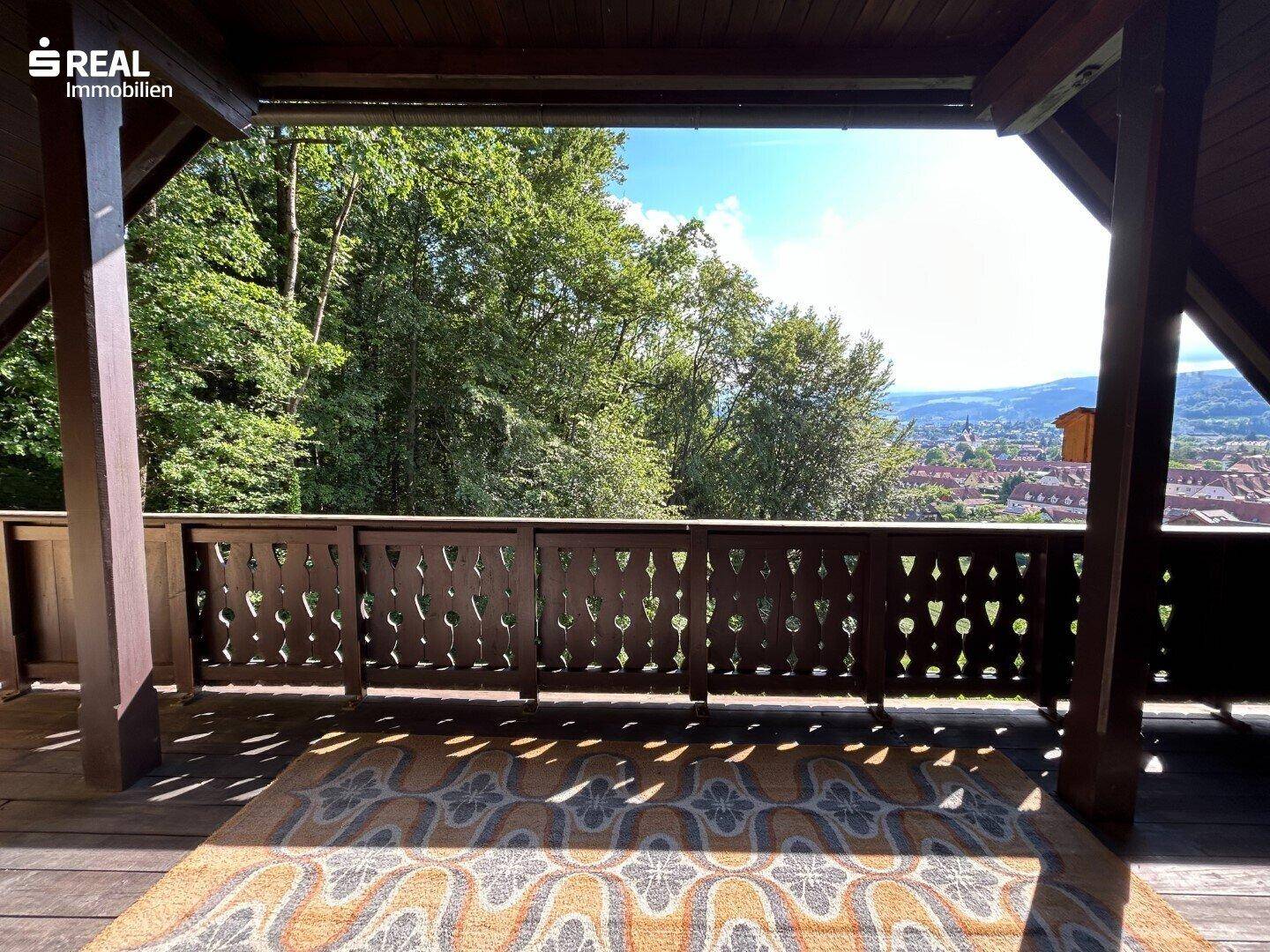 Überdachter Balkon mit Holzgeländer und Teppich, bietet einen schönen Blick auf Bäume und die Umgebung.
