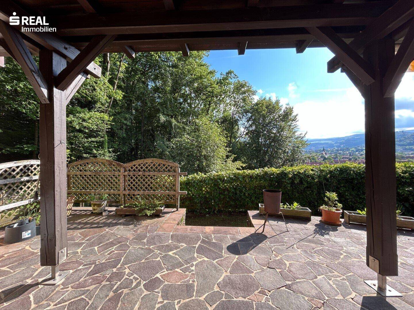 Überdachte Terrasse mit Steinplattenboden und Holzpergola, umgeben von üppigem Grün.