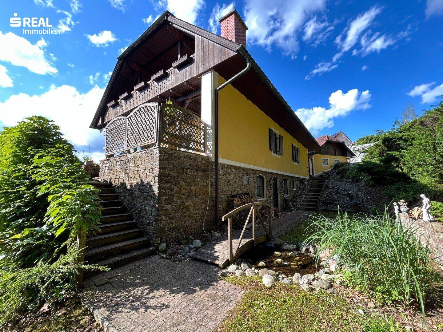 Außenansicht des Hauses von unten mit Steinfundament, Treppen, kleinem Teich und üppigem Garten.