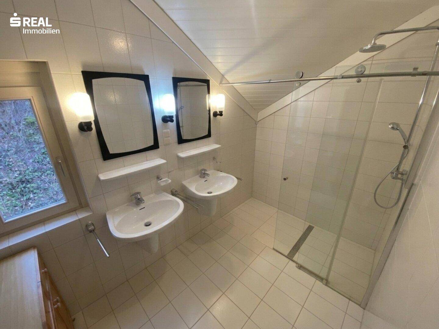 Helles Badezimmer mit zwei Waschbecken, großer Dusche und Dachschräge für natürliches Licht.