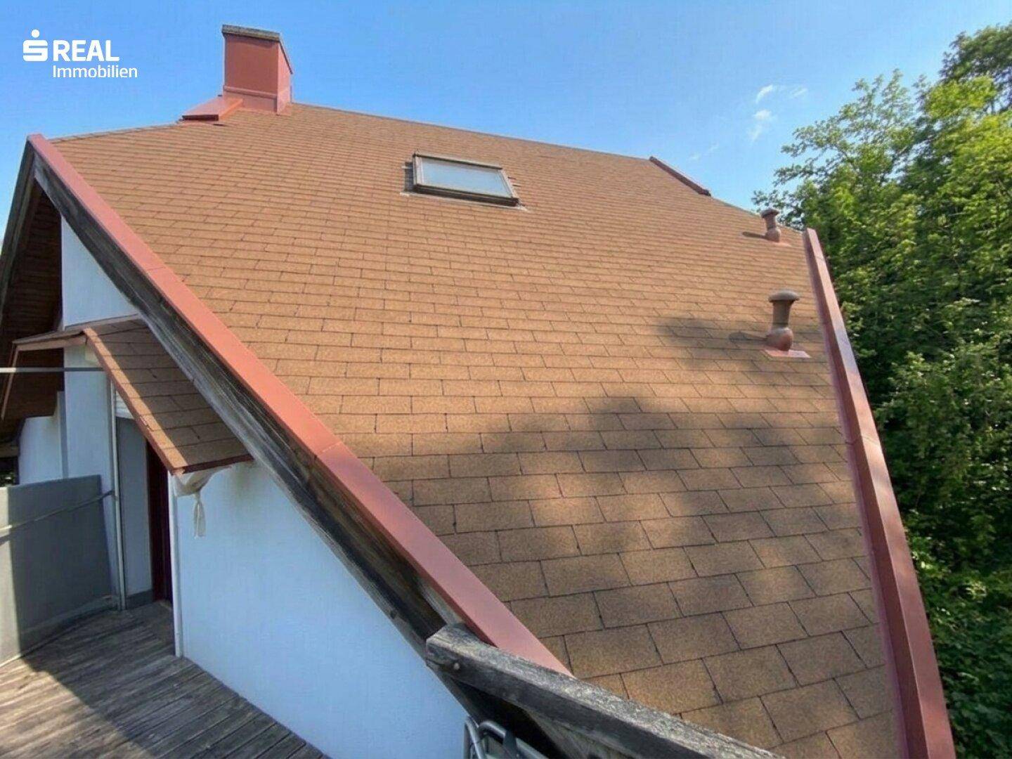 Braunes Ziegeldach mit Dachfenster und sichtbaren Dachrinnen unter blauem Himmel.