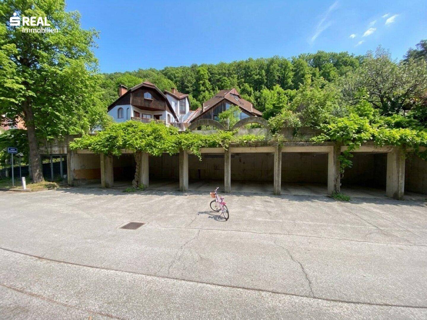 Außenparkplatz mit mehreren Garagen und üppiger Begrünung im Hintergrund der Gebäude.