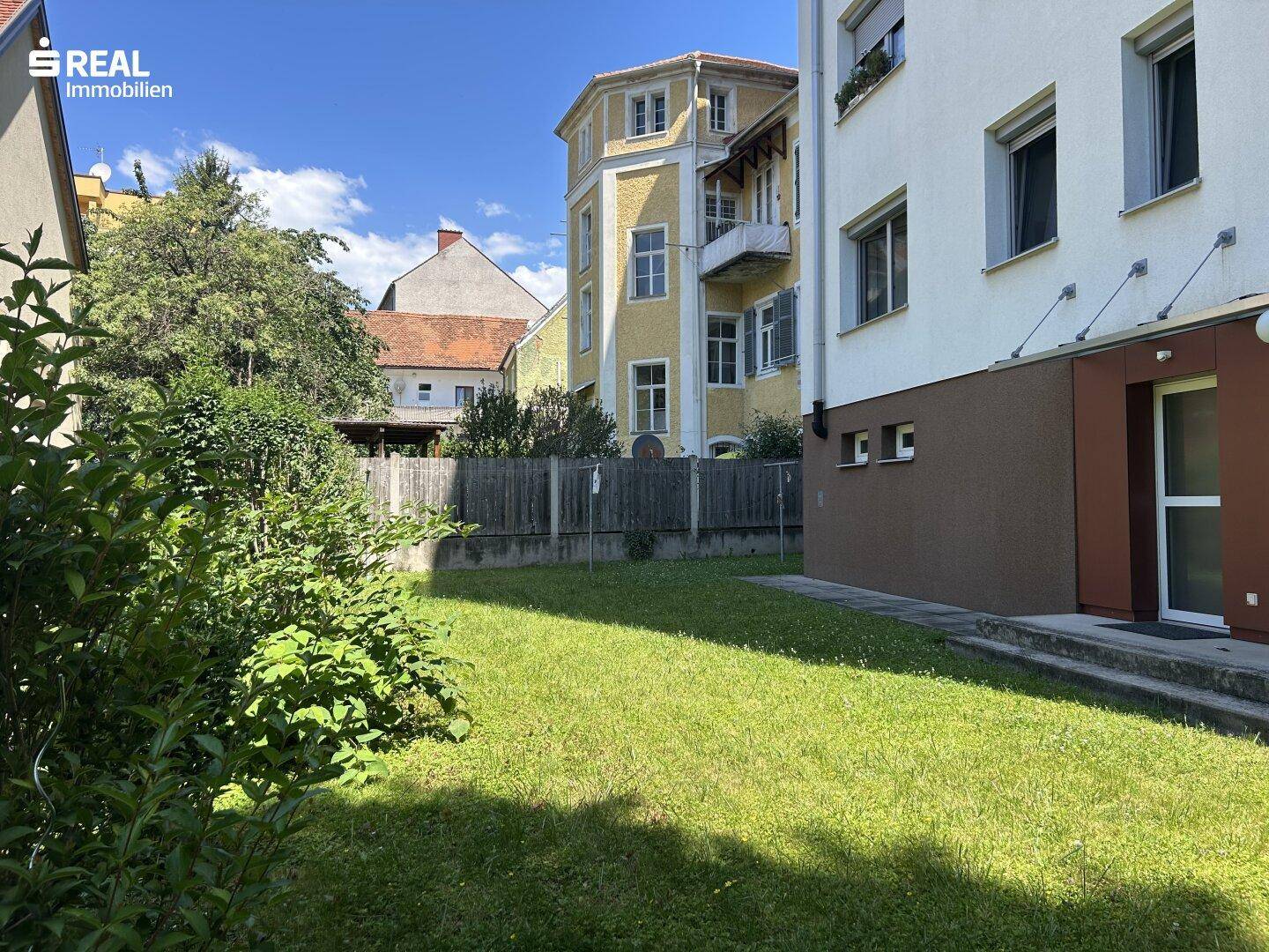 Grüner Gartenbereich mit Rasenfläche und Bäumen hinter einem Wohngebäude.