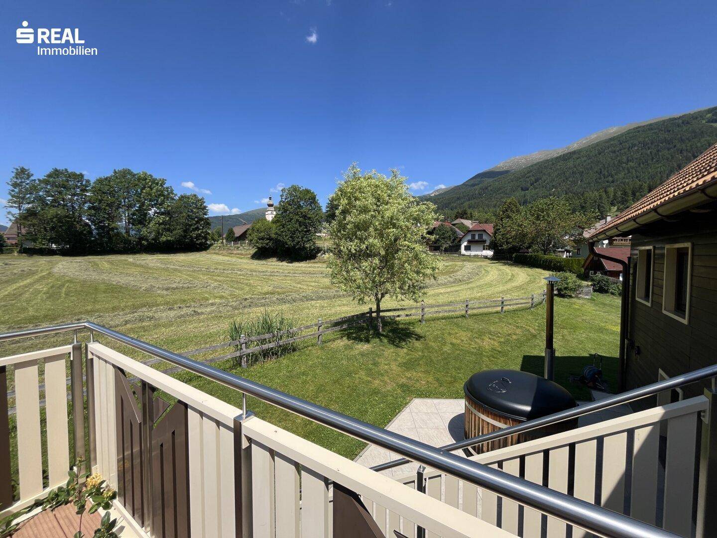 Blick von der Terrasse auf grüne Wiesen und Berge mit einem Whirlpool unter blauem Himmel.