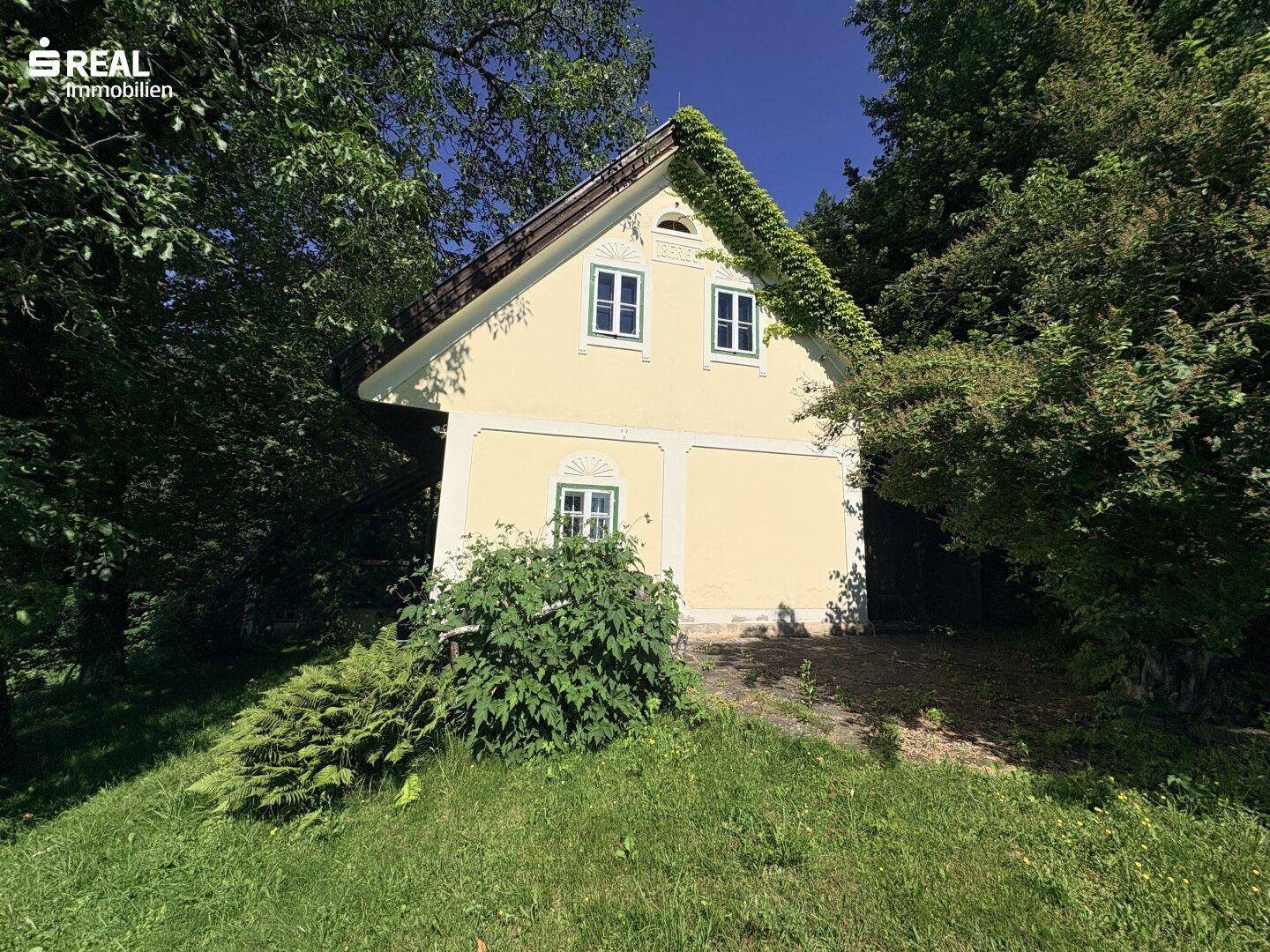 Gelbe Hausfassade mit grünen Fensterläden und Giebel, umgeben von dichtem Baumbestand.
