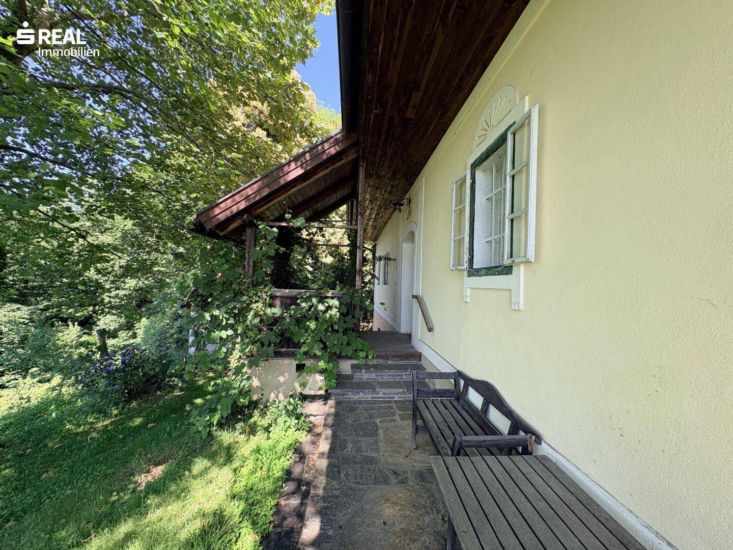 Überdachter Eingangsbereich mit Holzbänken und grünen Fensterläden, umgeben von Natur.