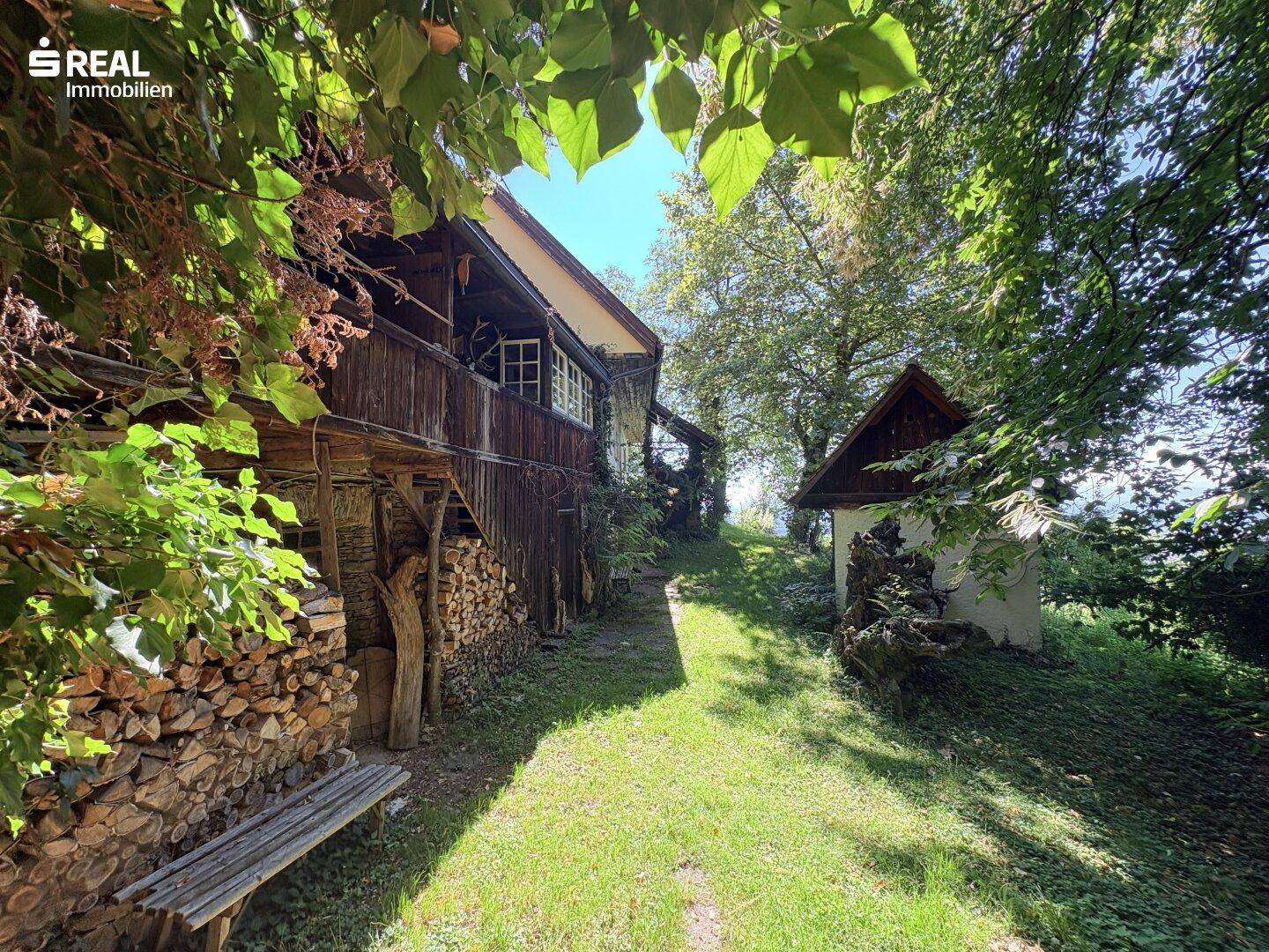 Rustikales Anwesen mit Holzstapeln und einem kleinen Nebengebäude, umgeben von üppigem Grün.
