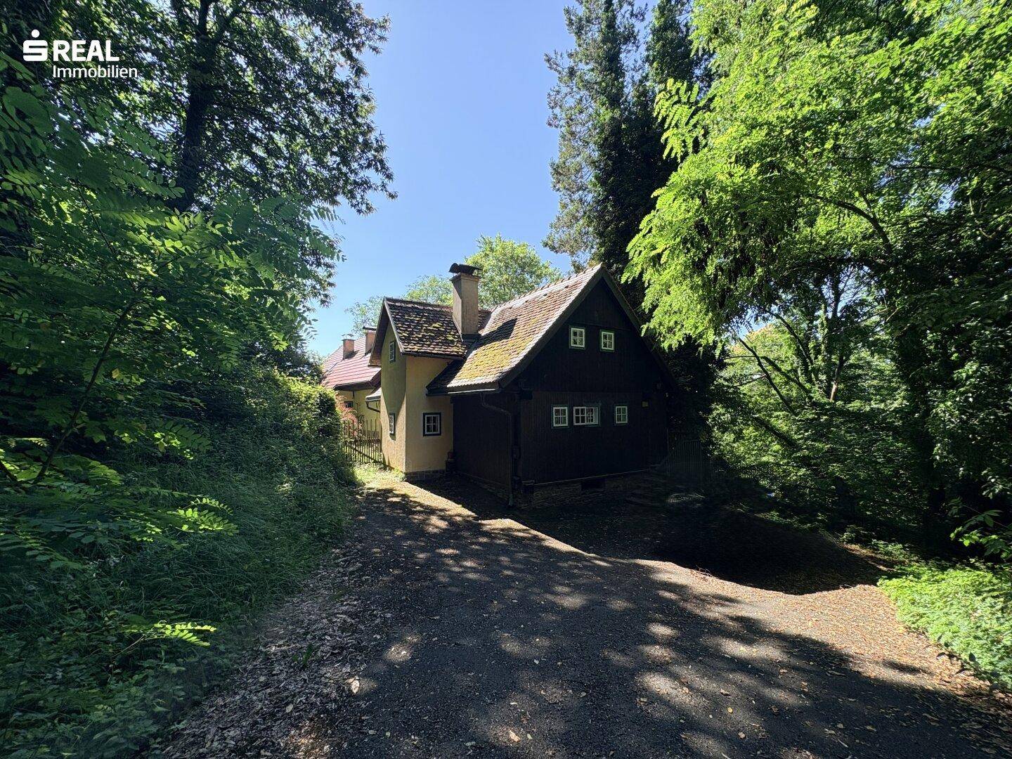 Landhaus mit Holzelementen und Schornstein, umgeben von dichtem Wald und einem Schotterweg.