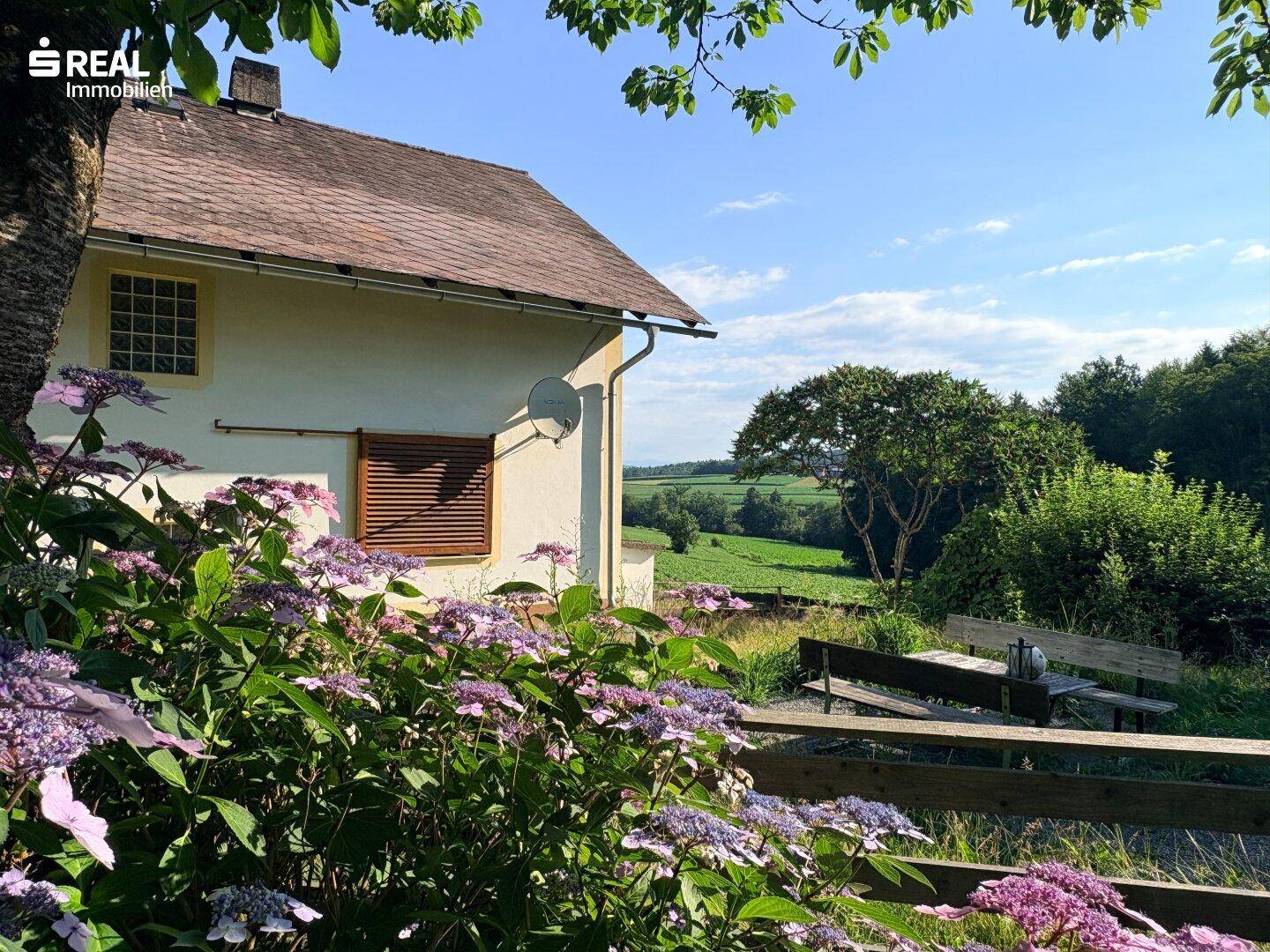 Seitenansicht des Hauses mit blühenden Hortensien, Holzzaun und weitem Blick über die Landschaft.