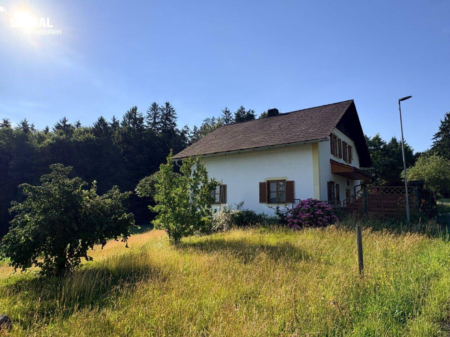Landhaus mit Satteldach und heller Fassade, umgeben von Wiesen und Wald unter blauem Himmel.