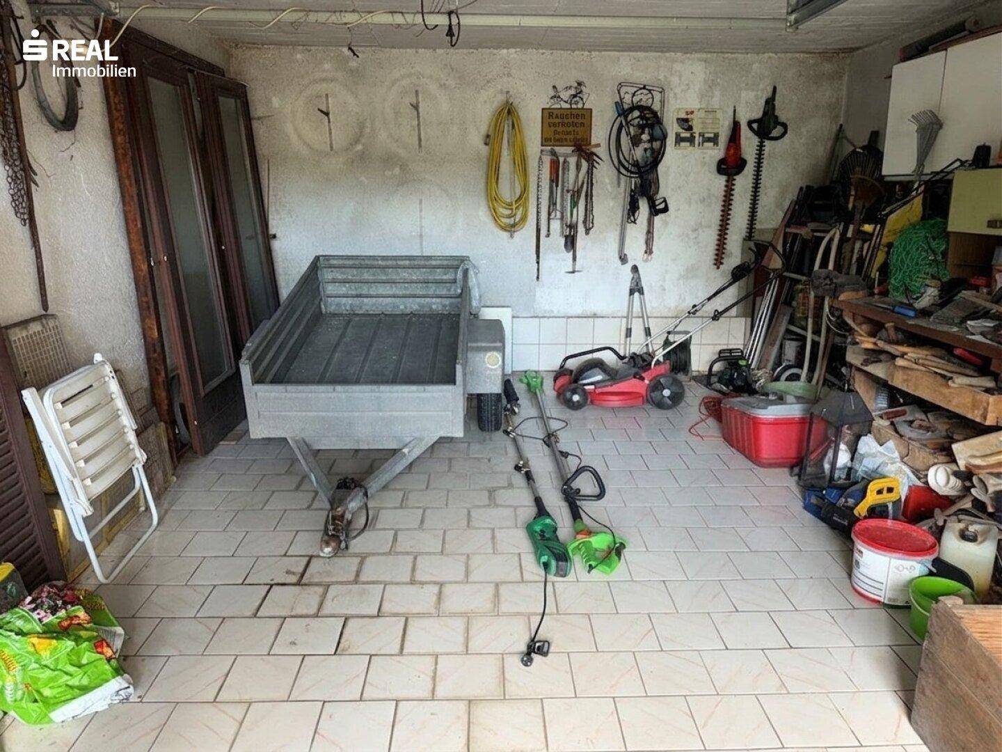 Garage oder Abstellraum mit Anhänger, Gartengeräten und Werkzeugen.
