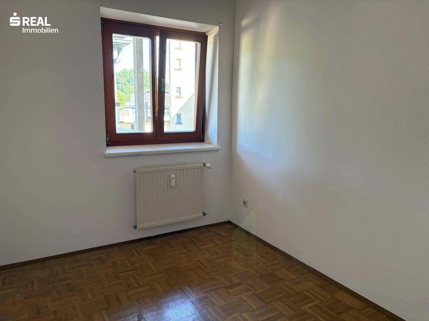 Helles Schlafzimmer mit Parkettboden und Fenster, das viel Tageslicht hereinlässt.