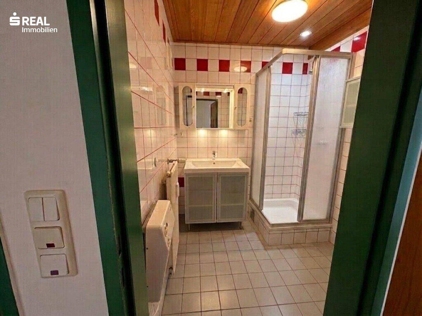 Badezimmer mit Dusche, Waschbecken und Spiegelschrank, gefliest in Weiß und Rot.