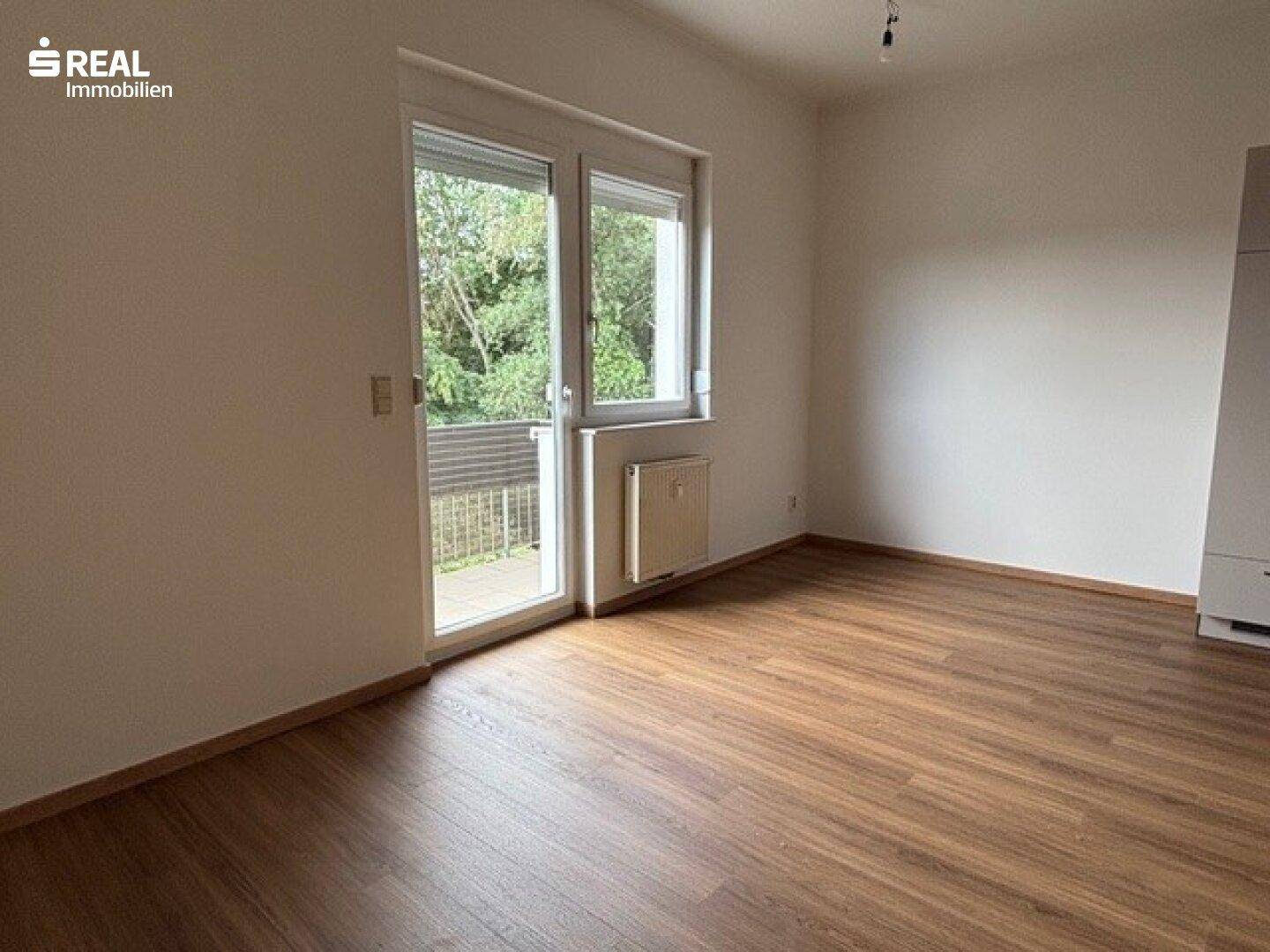 Heller Raum mit Laminatboden und Zugang zu einem Balkon, bietet Blick ins Grüne.