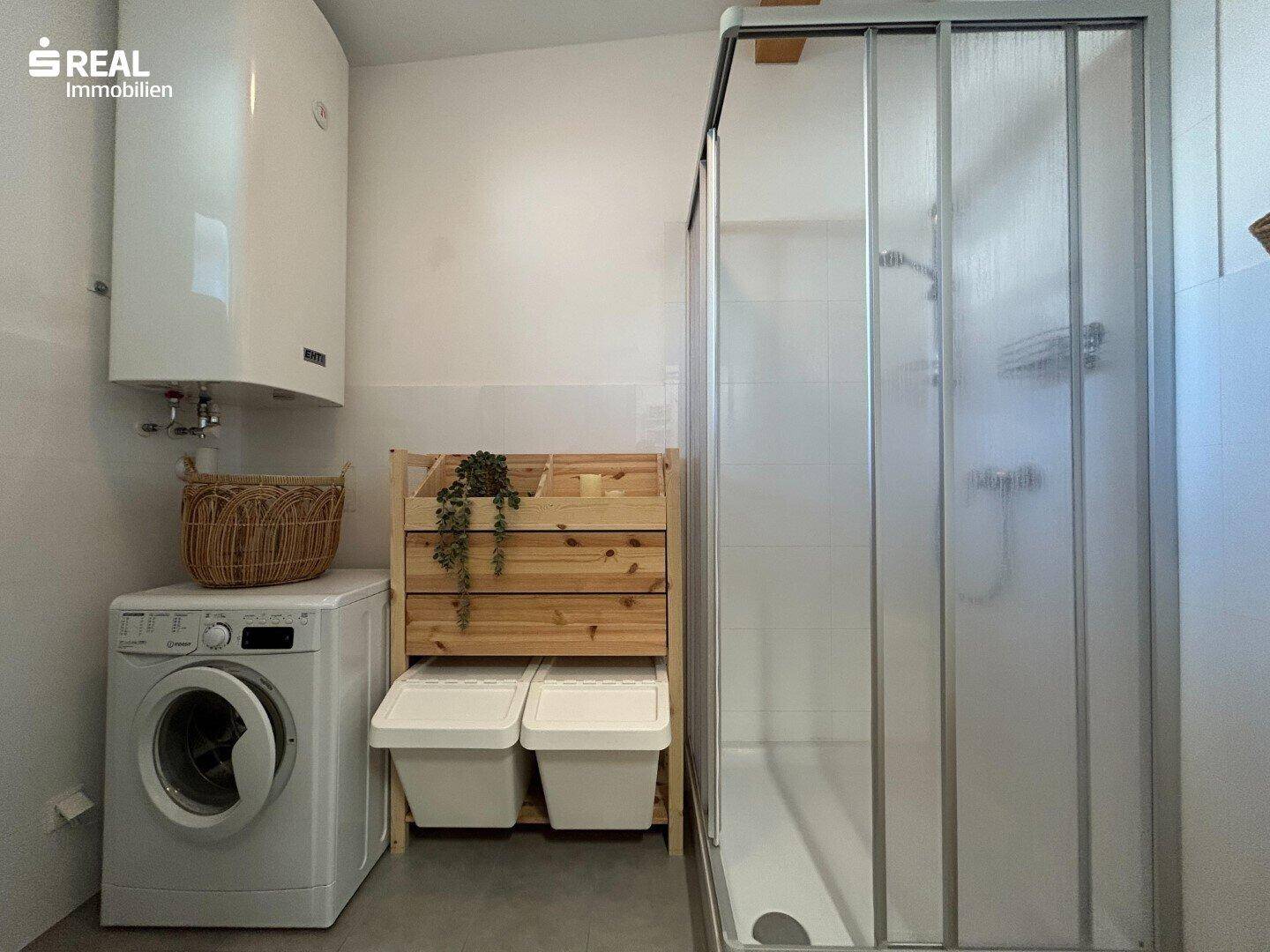Badezimmer mit Duschkabine, Waschmaschine und Warmwasserboiler an der Wand.