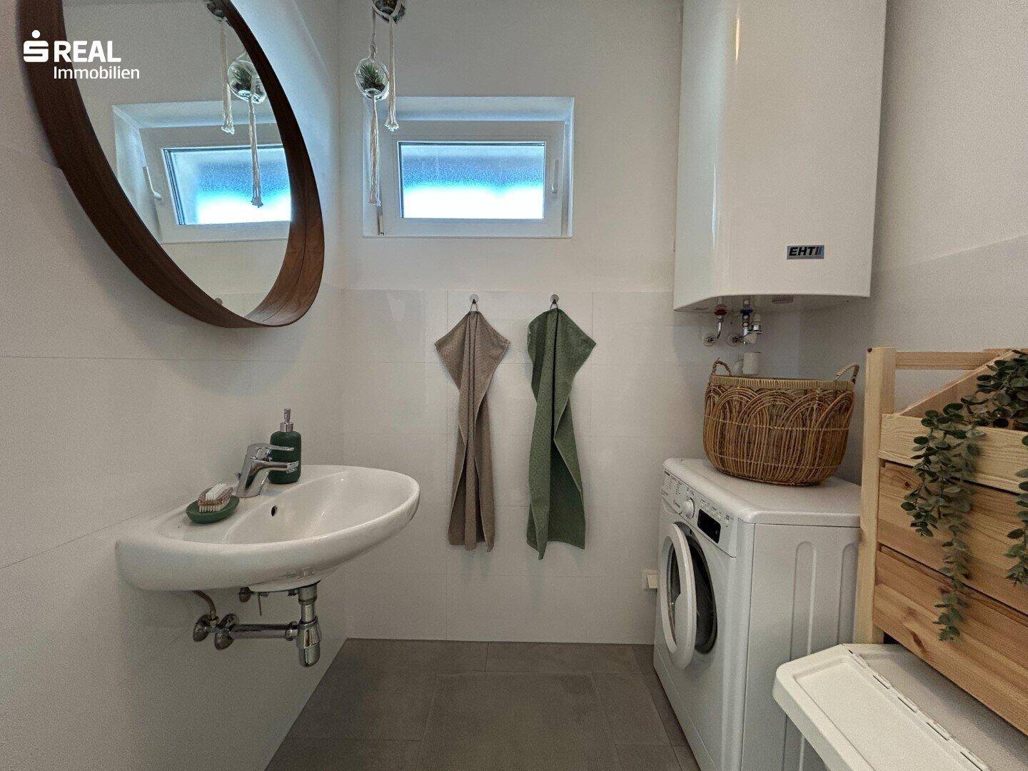 Helles Badezimmer mit Waschbecken, Spiegel, Waschmaschine und zwei kleinen Fenstern.