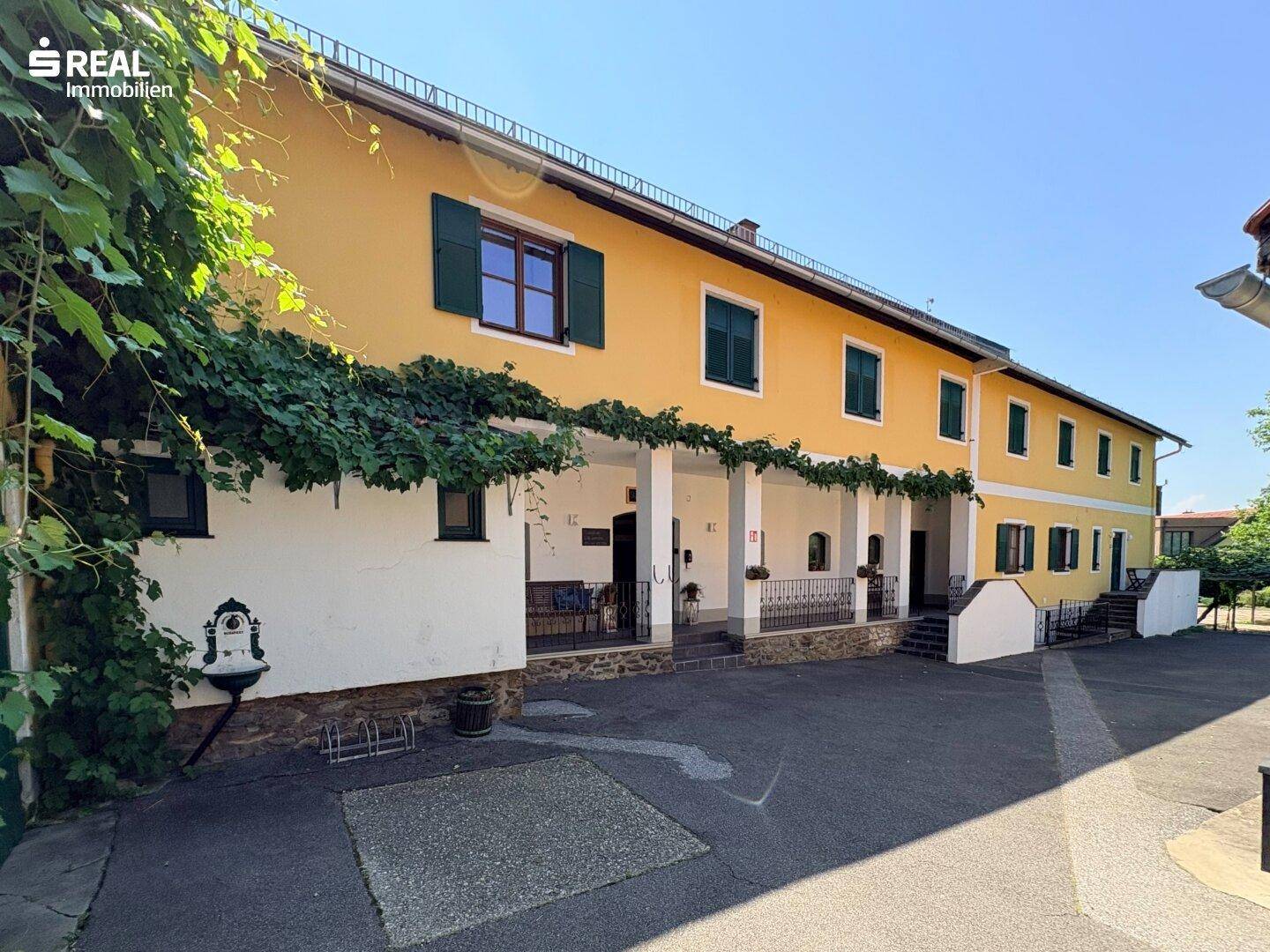 Langes gelbes Gebäude mit überdachter Terrasse, grünen Fensterläden und einem gepflasterten Vorplatz.