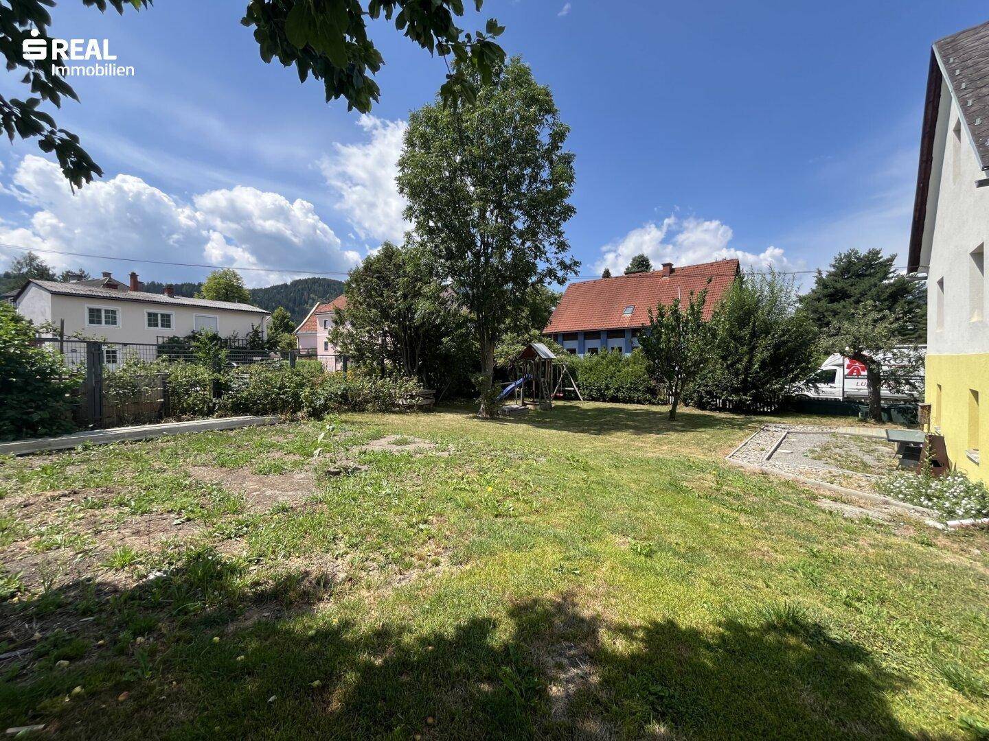 Gepflegter Gartenbereich mit Rasenfläche, großem Baum und Kinderspielplatz im Hintergrund.