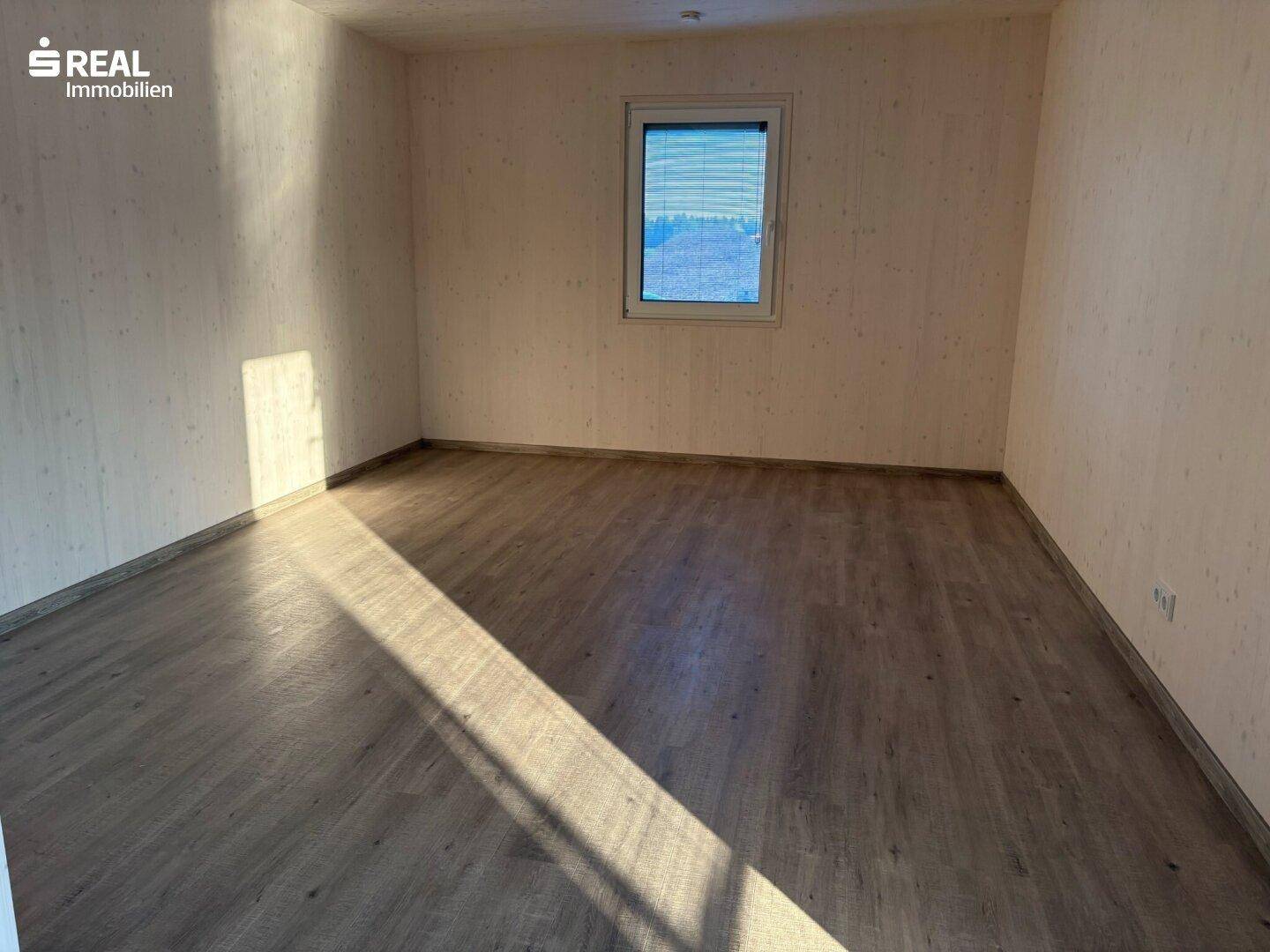 Leerer Raum mit Laminatboden und einem Fenster, das natürliches Licht hereinlässt.