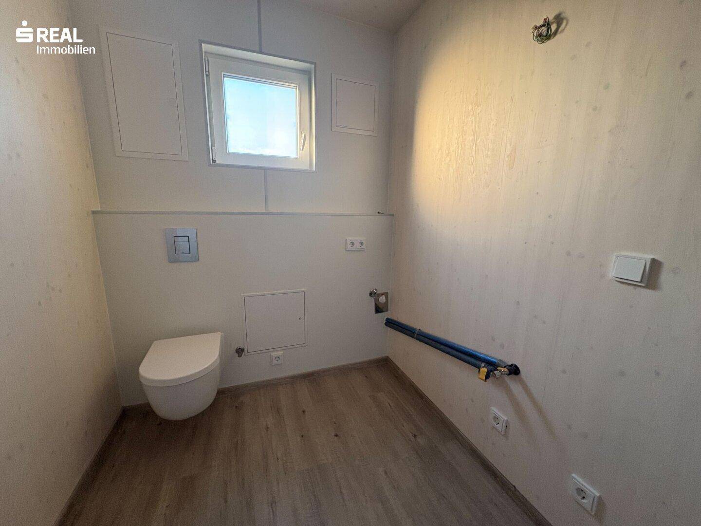 Kleines WC mit wandhängender Toilette, Fenster und vorbereiteten Anschlüssen für ein Waschbecken.