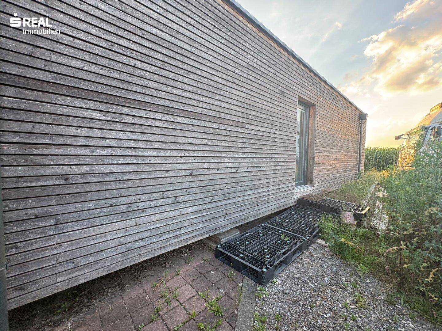 Seitenansicht eines modernen Holzgebäudes mit schmalem Fenster und ländlicher Umgebung im Hintergrund.
