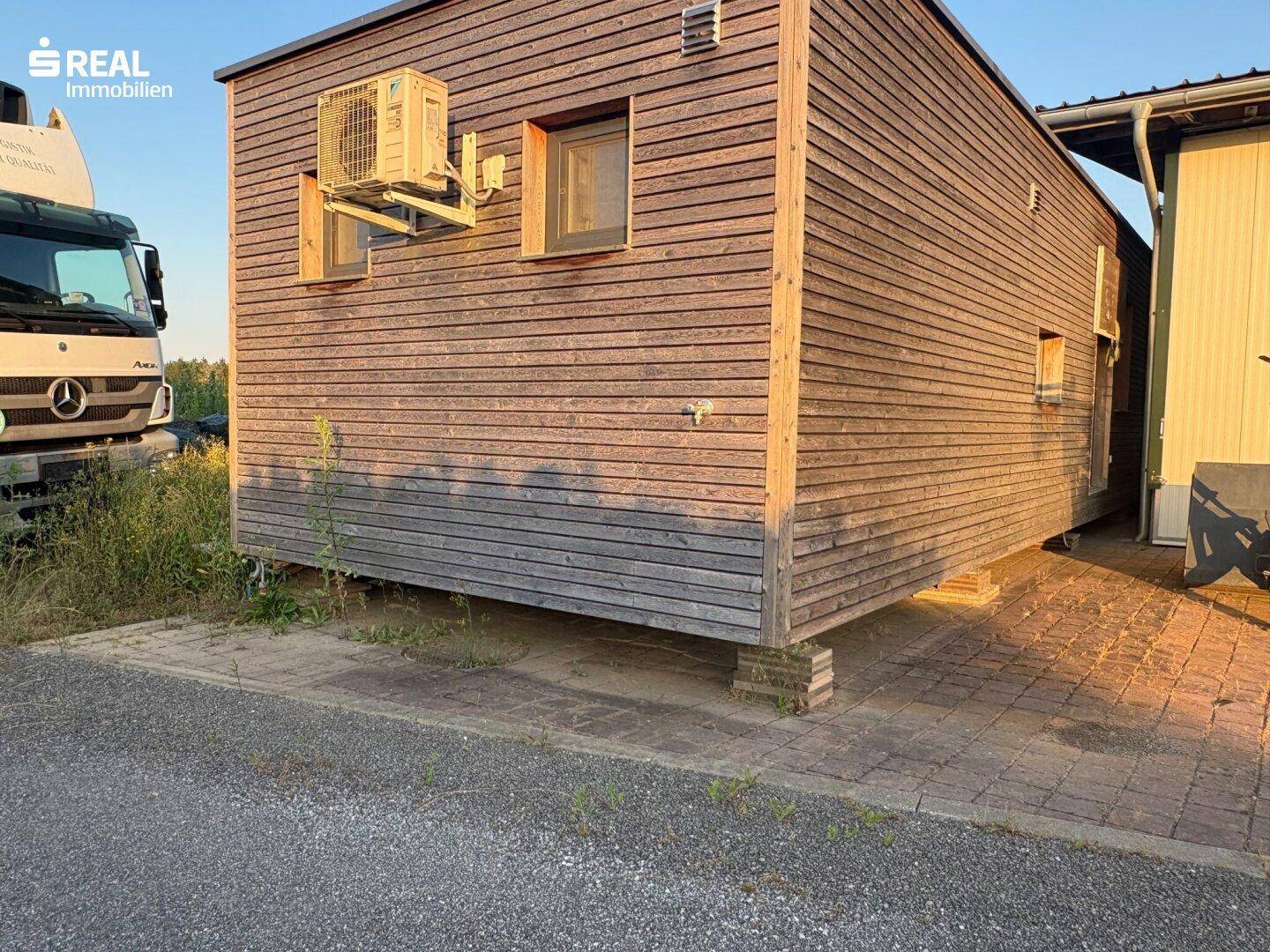 Holzverkleidetes Gebäude mit Klimaanlage an der Fassade und gepflastertem Vorplatz im Freien.