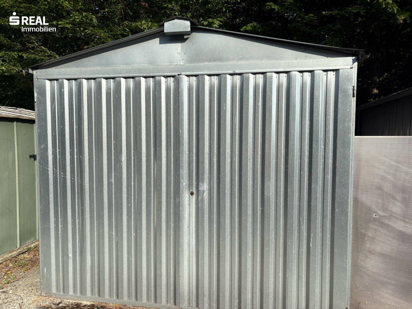 Einfache Garage aus Wellblech mit einem Satteldach, umgeben von Bäumen.