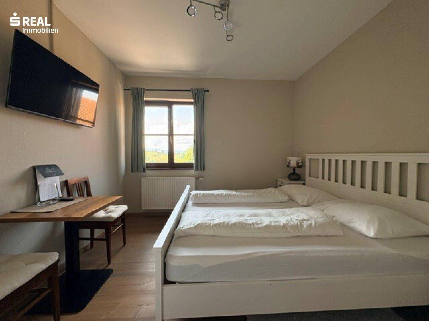 Helles Schlafzimmer mit Doppelbett, kleinem Tisch und Blick ins Grüne durch das Fenster.
