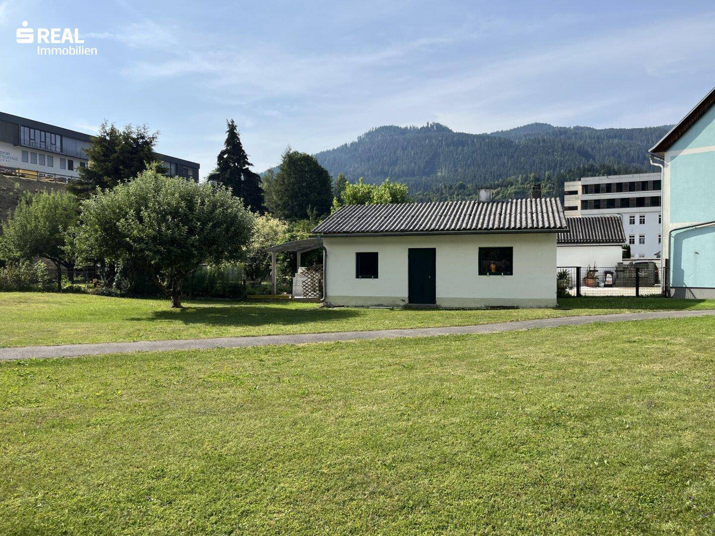 Kleines weißes Gebäude mit Rasenfläche und Bergblick unter strahlend blauem Himmel.