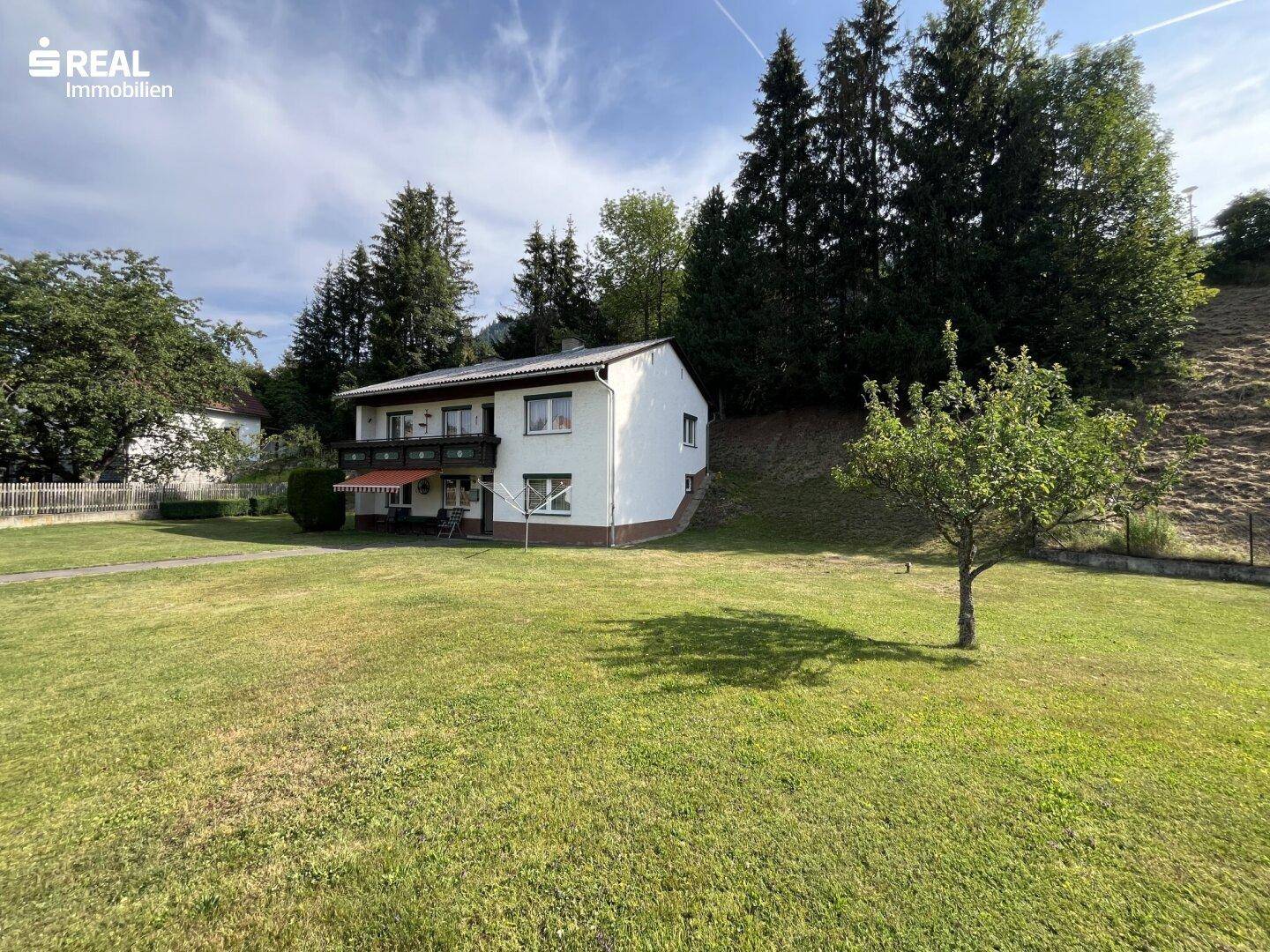 Das Haus mit großem Rasen und bewaldetem Hang im Hintergrund unter blauem Himmel.