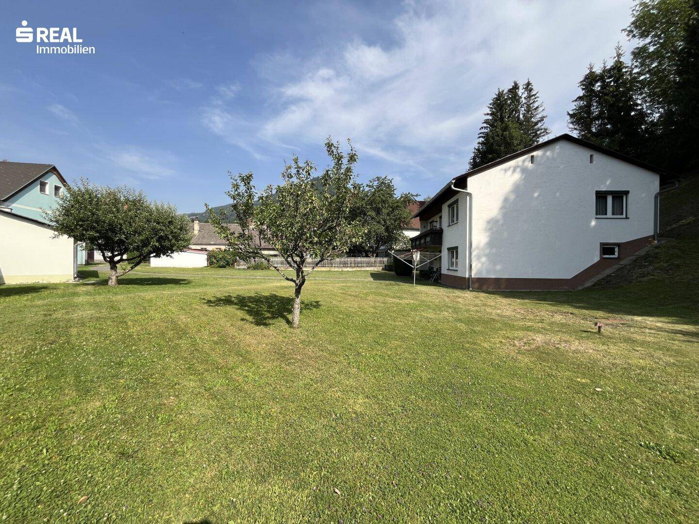 Seitenansicht des Hauses mit großem Rasen und Bäumen unter blauem Himmel.