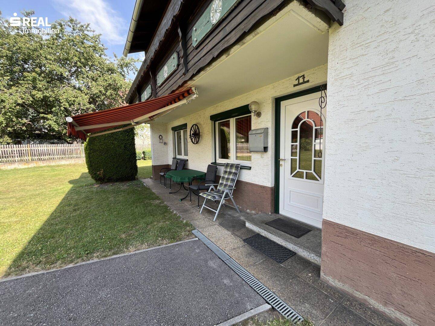 Eingangsbereich des Hauses mit kleiner Terrasse und Markise, umgeben von Grün.