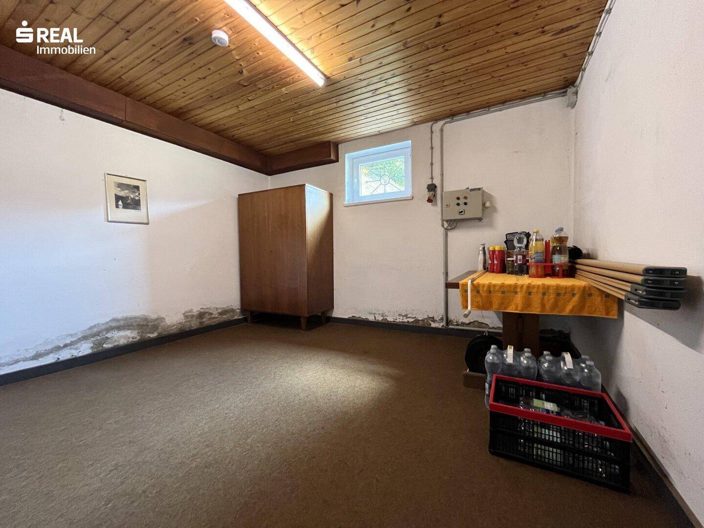 Kellerraum mit Holzdecke, Teppichboden und kleinem Fenster, genutzt als Abstellfläche.