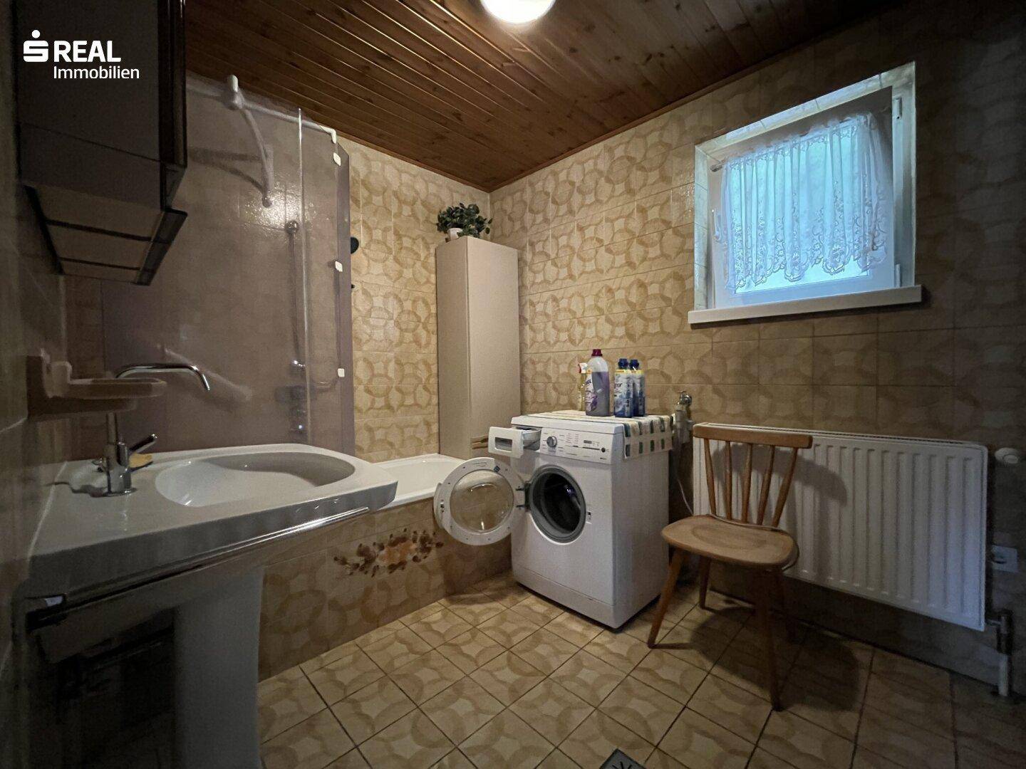 Badezimmer mit Badewanne, Waschmaschine und Fenster, gefliest im 80er-Jahre-Stil.