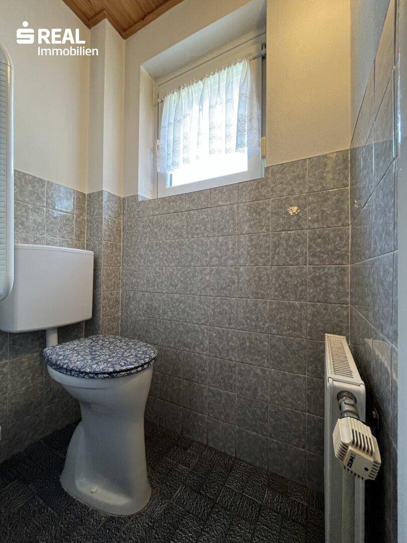 Kleines WC mit grauen Fliesen, Fenster und Holzdecke, ausgestattet mit Heizkörper.