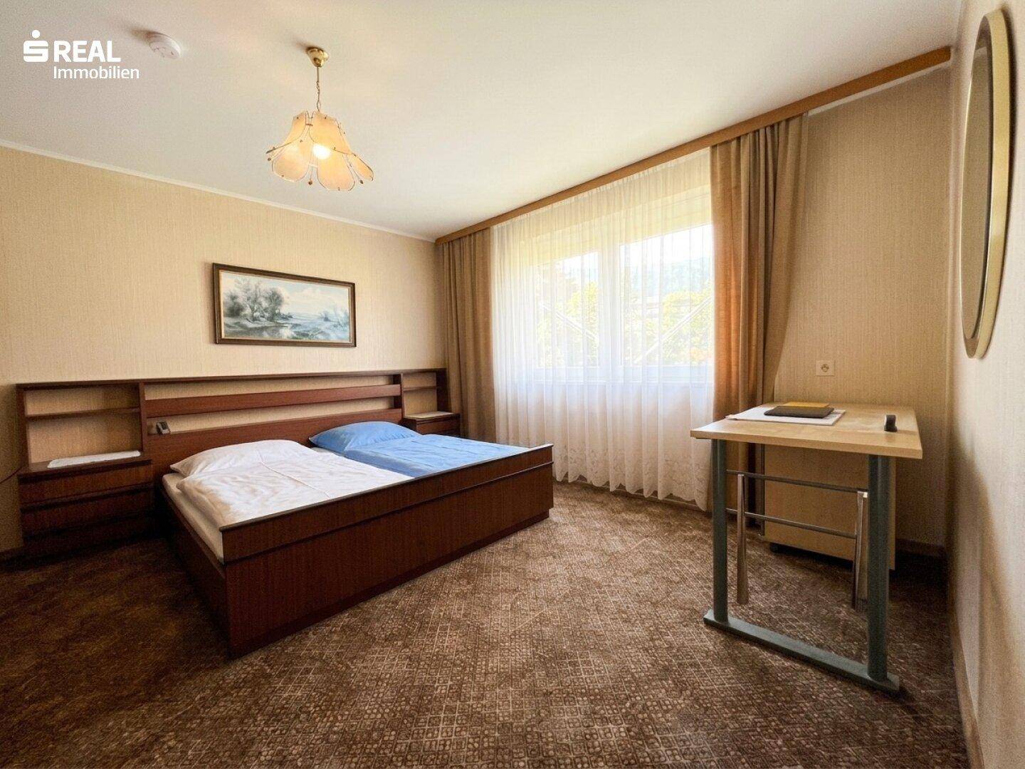 Schlafzimmer mit Doppelbett, Teppichboden und großem Fenster mit Vorhängen.