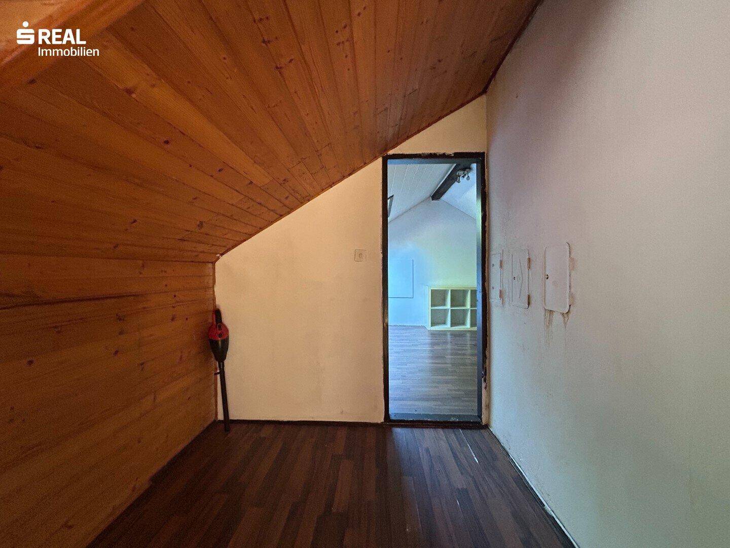 Dachgeschossraum mit Holzdecke, Laminatboden und Zugang zu einem weiteren Bereich.