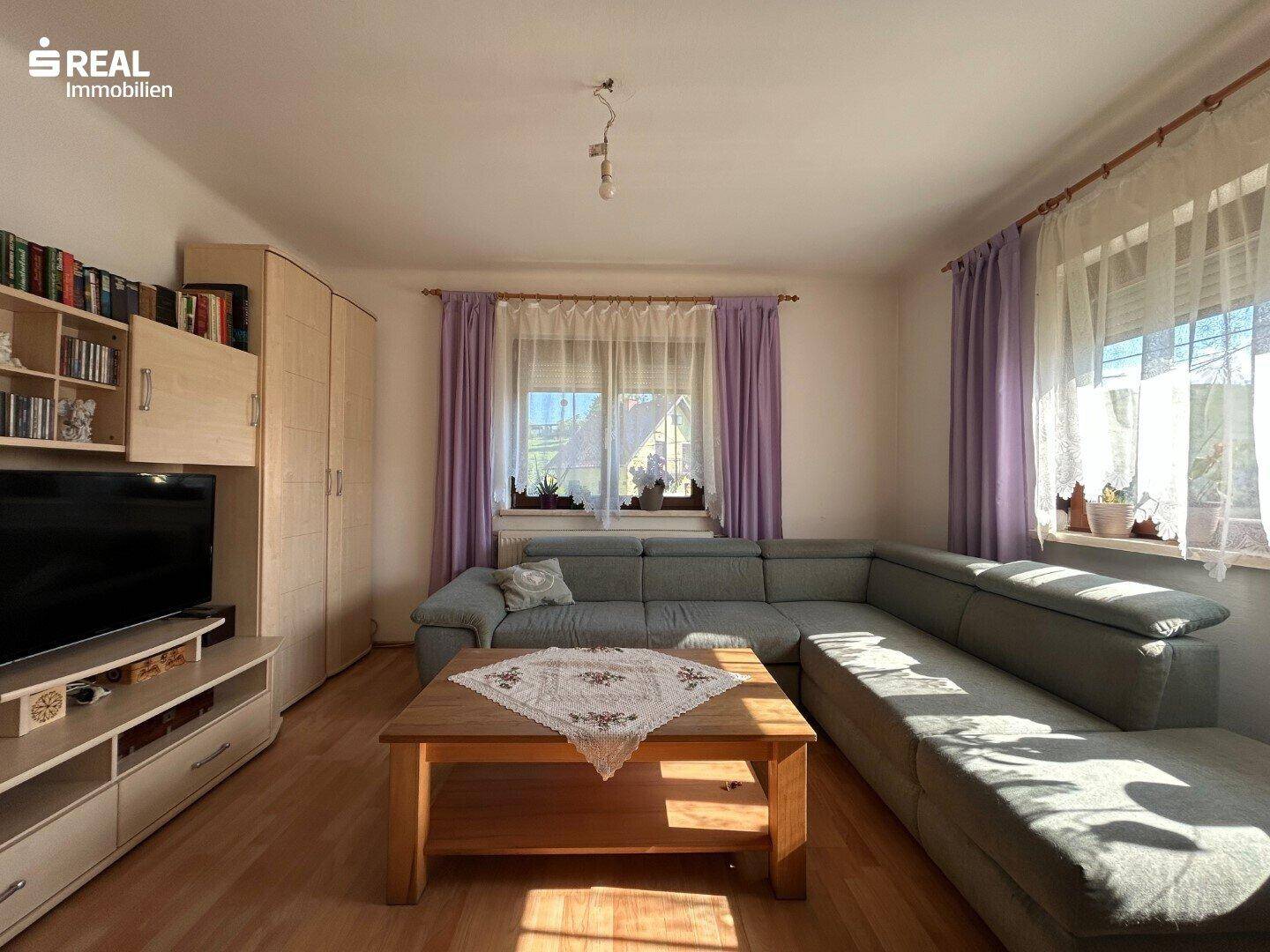 Gemütliches Wohnzimmer mit Ecksofa, Holzboden und viel Tageslicht durch die Fenster.