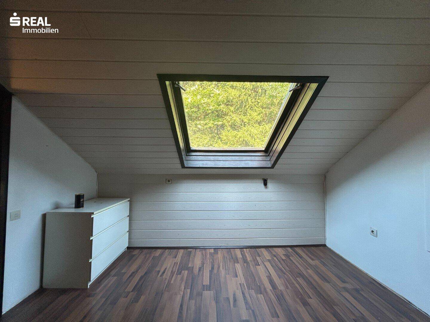 Dachgeschossraum mit großem Dachfenster, Laminatboden und weißer Kommode.