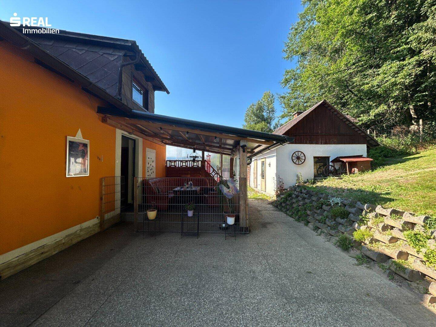 Überdachte Terrasse neben dem Haus mit Blick auf ein Nebengebäude und viel Grün.