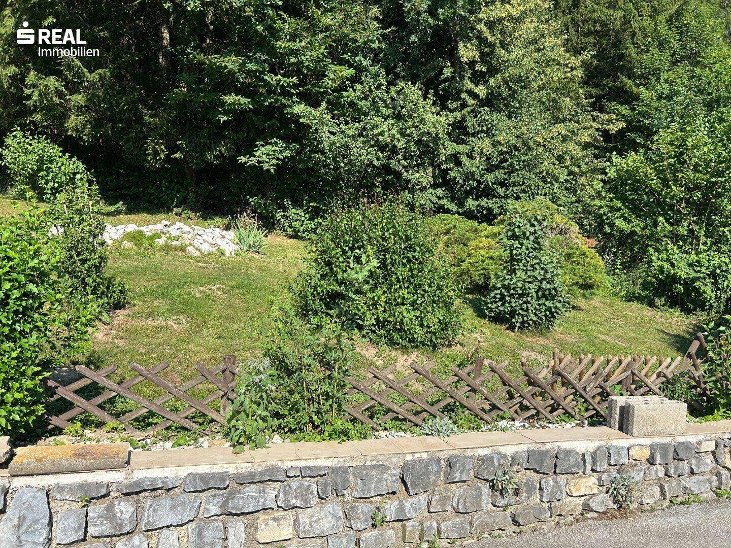 Hanglage Garten mit üppigem Grün, Bäumen und einem rustikalen Holzzaun auf einer Steinmauer.