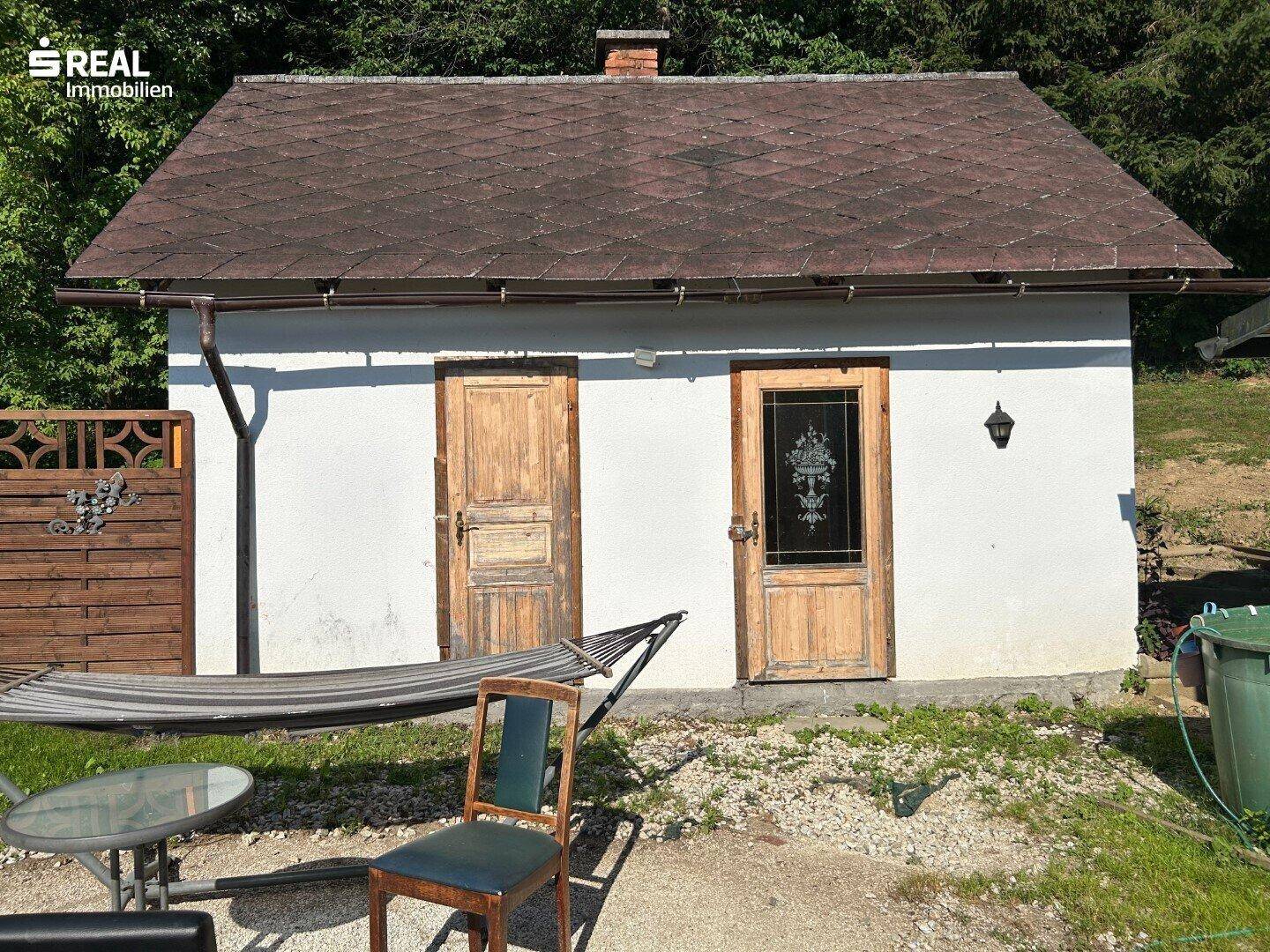 Kleines Nebengebäude mit zwei Holztüren, einer Hängematte und Gartenmöbeln davor.