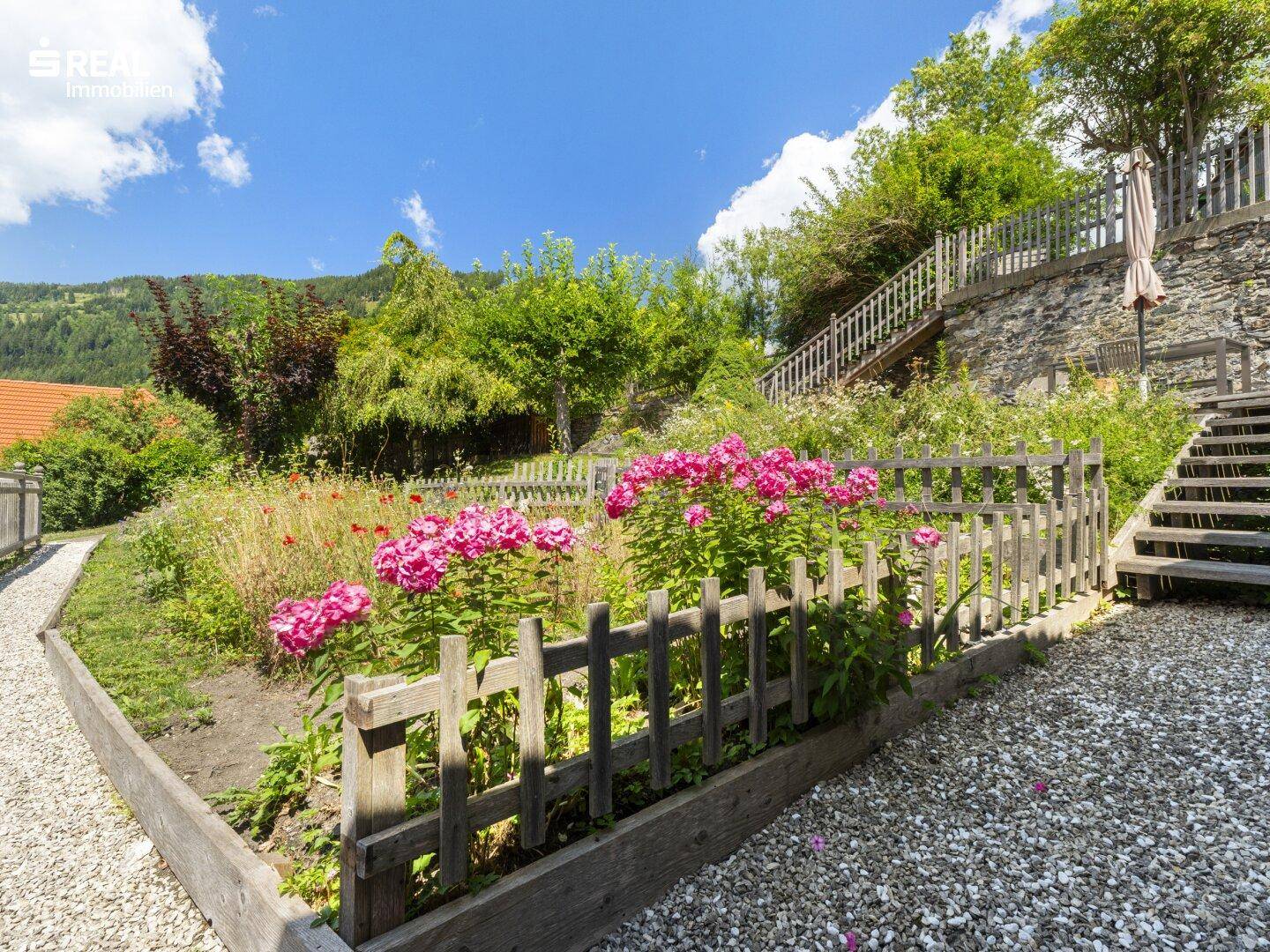 Malerischer Gartenbereich mit Blumenbeeten, Holzzaun und Kieswegen, umgeben von üppigem Grün.