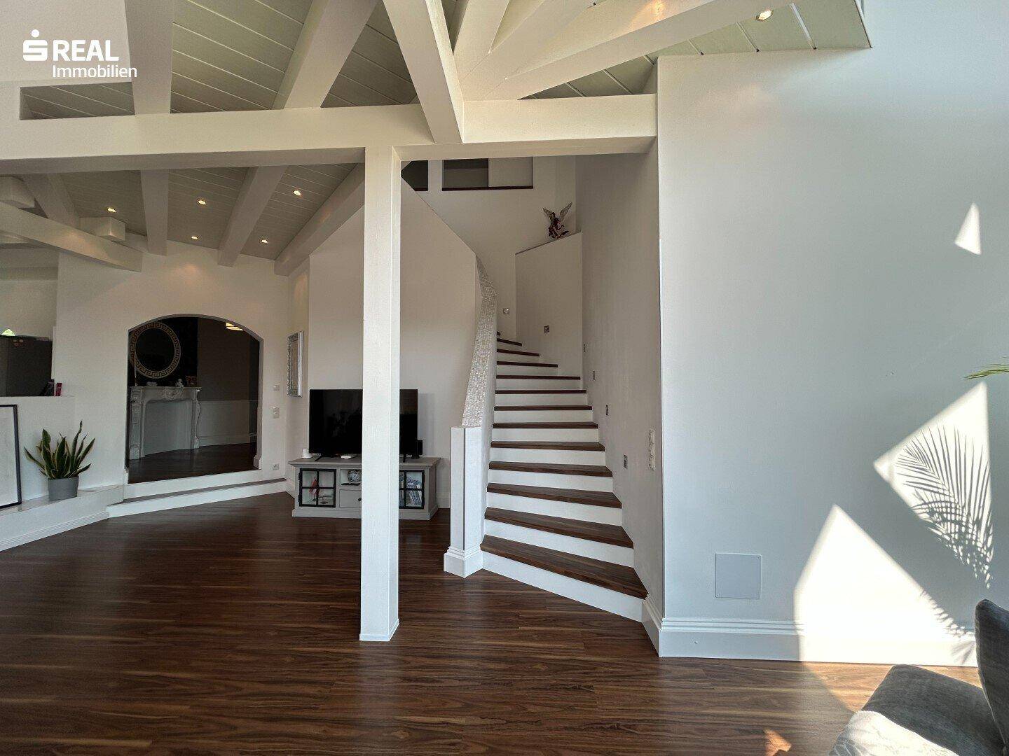 Heller Wohnbereich mit offener Treppe, hohen Decken und dunklem Holzboden.