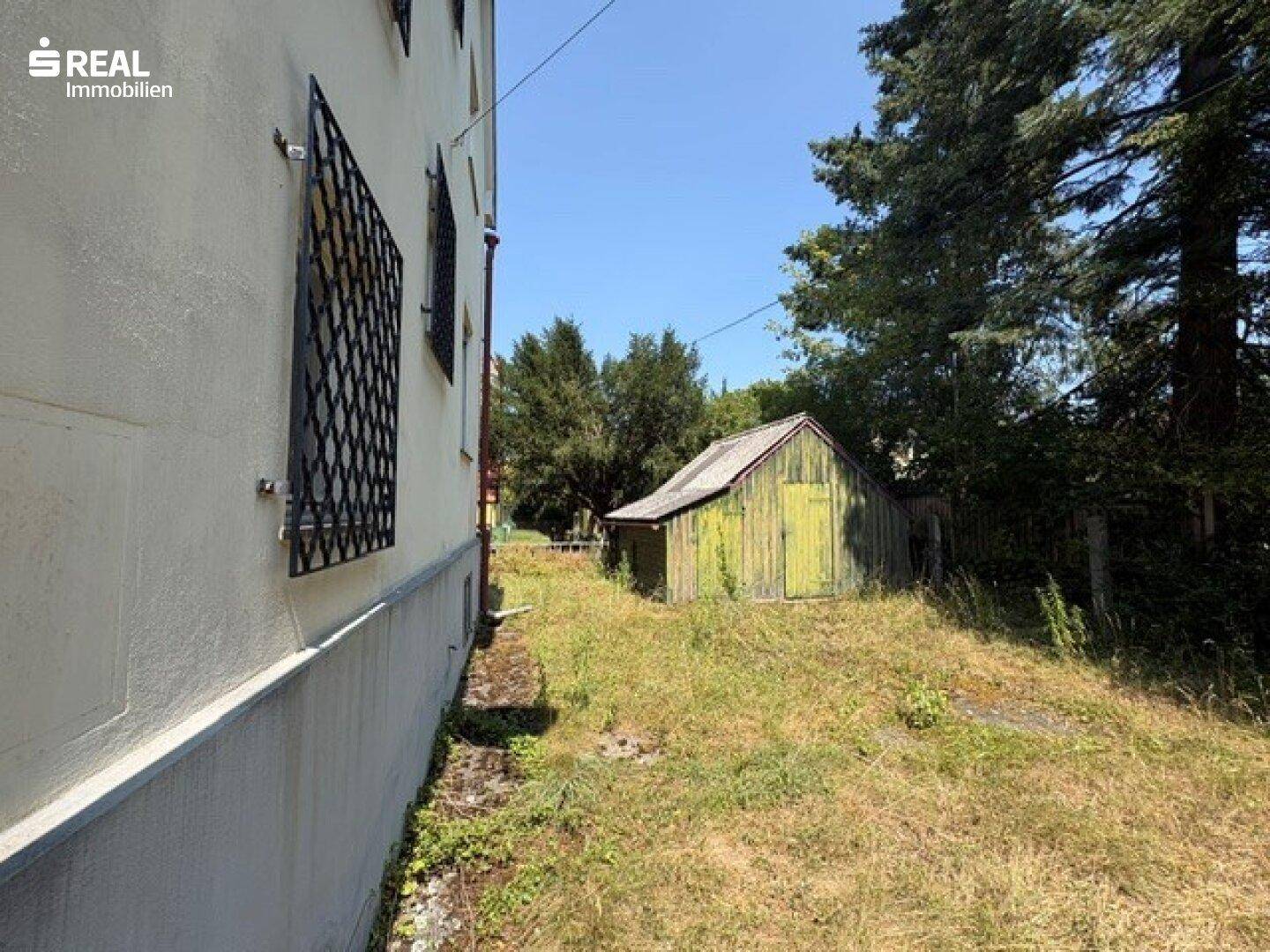 Seitenansicht des Hauses mit vergitterten Fenstern und einem rustikalen Gartenschuppen im Grünen.